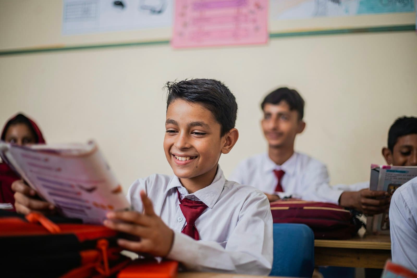 A smiling student in school uniform reading a book at his desk, classmates behind him