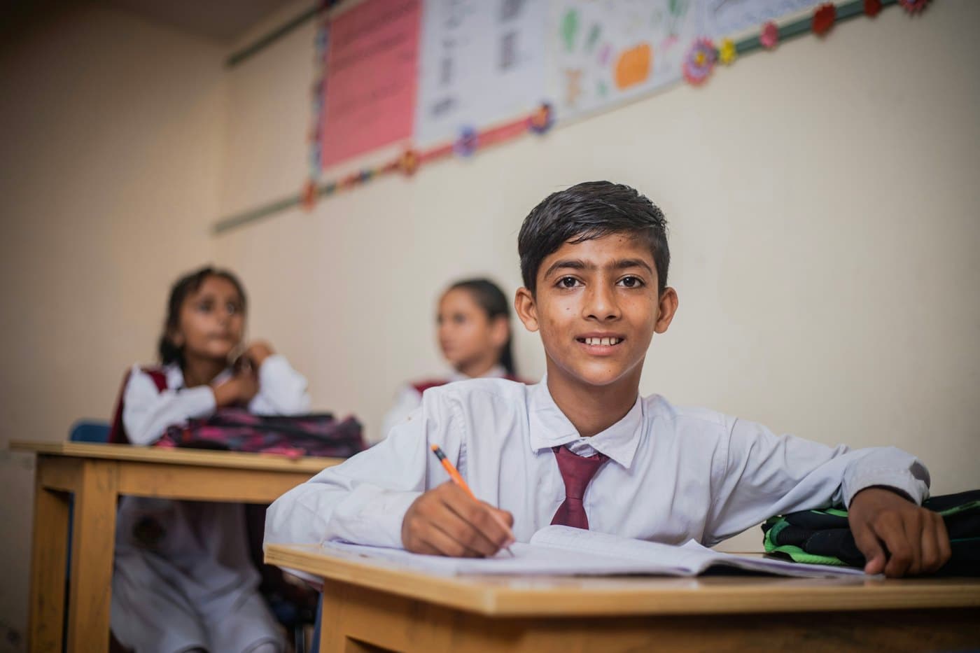 A student in a white shirt and red tie writing in his exercise book at his desk